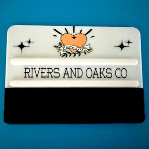 Puede incluir: Una espátula blanca con borde de fieltro negro, con un diseño de tatuaje de corazón con las palabras "Pure Love" y el texto "Rivers and Oaks Co." sobre un fondo azul. Incluye gráficos de ráfagas de estrellas y estrellas.