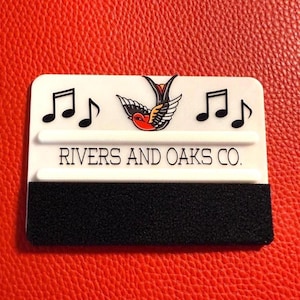 Puede incluir: Una espátula rectangular blanca con notas musicales negras y un diseño de tatuaje de pájaro rojo, negro y blanco. El texto "RIVERS AND OAKS CO." está impreso en la espátula. Una tira de espuma negra está en la parte inferior.