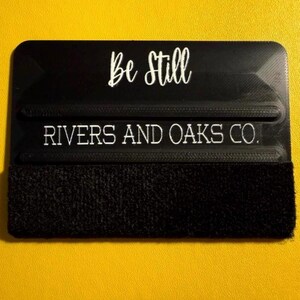 May include: A black squeegee with the words "Be Still" and "RIVERS AND OAKS CO." in white script. The bottom edge has a black, textured material. The background is a solid yellow.