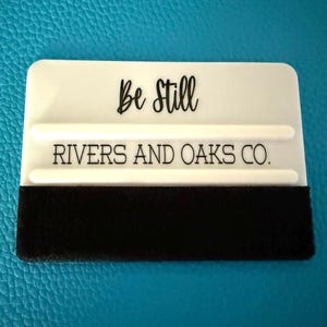 May include: A white squeegee with the words "Be Still" and "Rivers and Oaks Co." in black lettering. The bottom edge has a black felt strip. The squeegee is on a blue surface.