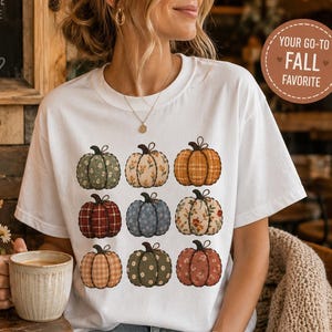 May include: A white short-sleeve t-shirt featuring a graphic print of nine pumpkins in a grid pattern. Each pumpkin displays a unique pattern, including floral, plaid, and solid designs in shades of orange, green, blue, and red.