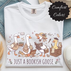 Puede incluir: Camiseta blanca con una ilustración caprichosa de cuatro gansos, cada uno leyendo o estudiando. Los gansos visten diferentes atuendos, con libros, gafas y un birrete. El texto "JUST A BOOKISH GOOSE" está impreso debajo.