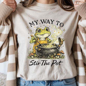 Puede incluir: Camiseta beige con una ilustración caprichosa de una rana. La rana, con un mono amarillo, revuelve un caldero burbujeante. El texto dice "MY WAY TO Stir The Pot" en una fuente lúdica, rodeado de estrellas y setas.