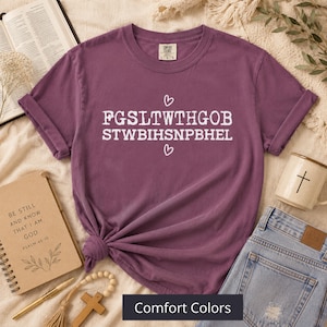 Könnte beinhalten: Ein pflaumenfarbenes Kurzarm-T-Shirt mit weißem Text, der FGSLTWTHGOB STWBIHSNPBHEL lautet, sowie einem kleinen Herzsymbol über und unter dem Text. Das T-Shirt hat einen Rundhalsausschnitt und eine lockere Passform, mit dem sichtbaren Etikett Comfort Colors.