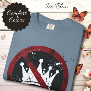 Puede incluir: Camiseta azul hielo con un diseño gráfico que presenta un círculo rojo con una línea que lo atraviesa, rodeando coronas blancas. La camiseta tiene las palabras "Comfort Colors" en una etiqueta negra. Mariposas y flores en el fondo.