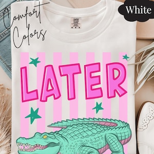 May include: White t-shirt with the text "LATER" in pink, a green alligator illustration, and pink and white striped background. The shirt also has green star graphics. The words "Comfort Colors" are written in cursive.