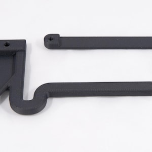 May include: Two matte black plastic components, possibly for a mechanical device, are displayed against a white background. One piece is rectangular with a curved extension and the other is a T-shaped piece. The pieces have a textured surface.