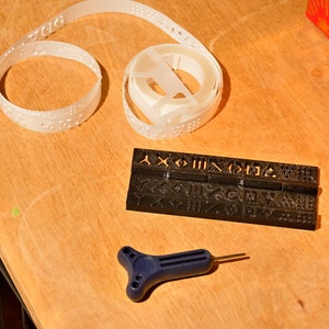 K&ouml;nnte beinhalten: Ein schwarzes Plastikwerkzeug mit erhabenen Buchstaben und Symbolen, eine wei&szlig;e Plastikrolle mit Braille-Punkten und ein blaues Plastikwerkzeug mit Griff und spitzer Spitze.