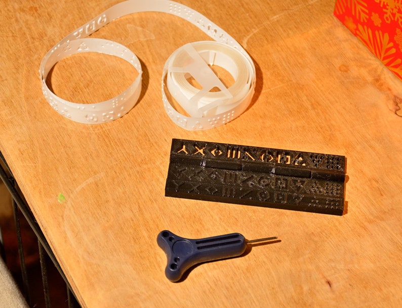 K&ouml;nnte beinhalten: Ein schwarzes Plastikwerkzeug mit erhabenen Buchstaben und Symbolen, eine wei&szlig;e Plastikrolle mit Braille-Punkten und ein blaues Plastikwerkzeug mit Griff und spitzer Spitze.