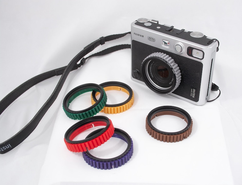 May include: A silver and black instant camera with a black leather strap and five colorful lens filters. The camera has the text "instax mini 9" printed on the side.