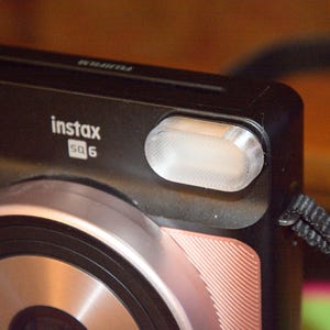 Könnte beinhalten: Nahaufnahme einer schwarzen Instax SQ6 Sofortbildkamera mit einem silbernen und roségoldenen Objektiv. Die Kamera hat einen eingebauten Blitz und einen schwarzen Riemen. Das Instax SQ6-Logo ist auf der Vorderseite sichtbar.