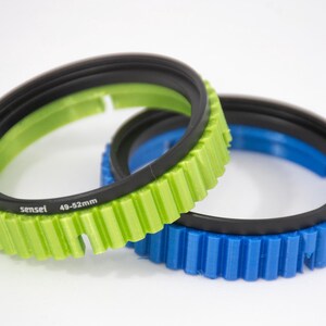 May include: Two step-up rings for camera lenses. The larger ring is green and has the text "sensel 49-52mm" printed on it. The smaller ring is blue.