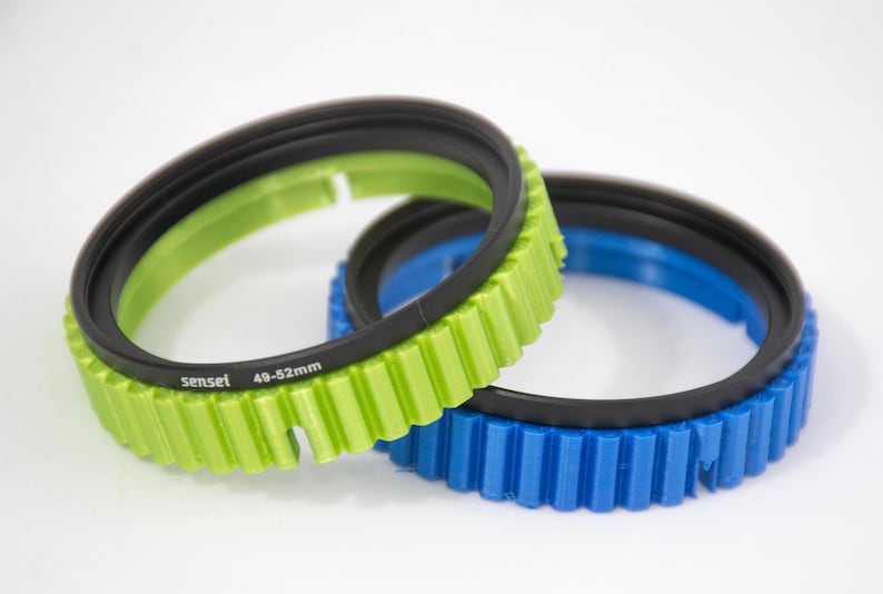 May include: Two step-up rings for camera lenses. The larger ring is green and has the text "sensel 49-52mm" printed on it. The smaller ring is blue.