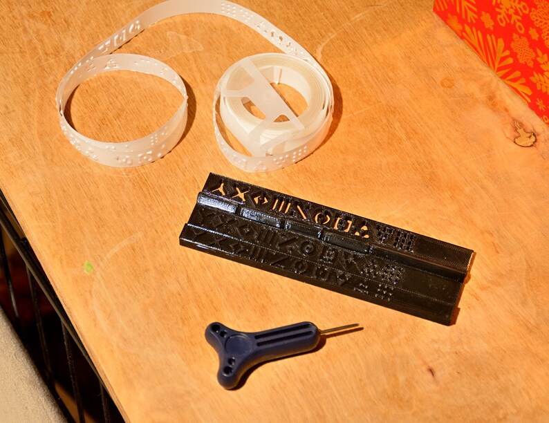 K&ouml;nnte beinhalten: Ein schwarzes Plastikwerkzeug mit erhabenen Buchstaben und Zahlen, eine wei&szlig;e Plastikspole mit Braille-Band und ein blaues Plastikwerkzeug mit T-f&ouml;rmigem Griff.