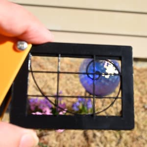 Puede incluir: Un marco negro con un patr&oacute;n de cuadr&iacute;cula y una l&iacute;nea curva en el interior. Una esfera de vidrio azul con una superficie reflectante es parcialmente visible en el marco. La esfera est&aacute; descansando sobre un lecho de mantillo marr&oacute;n y plantas verdes.