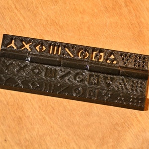 K&ouml;nnte beinhalten: Ein schwarzes Kunststoff-Scharnier mit einem dekorativen Muster aus Symbolen und Formen. Das Scharnier besteht aus zwei Teilen, die durch einen Metallstift verbunden sind.