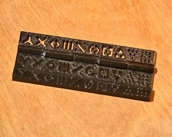 Könnte beinhalten: Ein schwarzes Kunststoff-Scharnier mit einem dekorativen Muster aus Symbolen und Formen. Das Scharnier besteht aus zwei Teilen, die durch einen Metallstift verbunden sind.