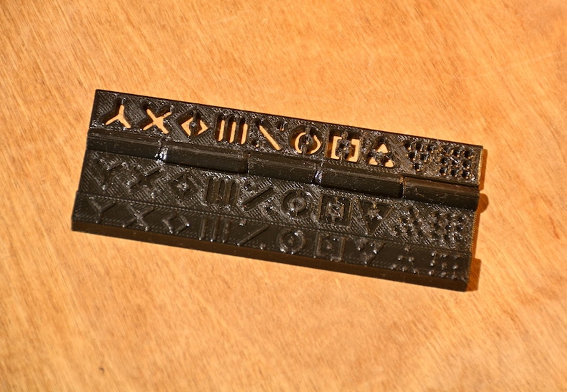 K&ouml;nnte beinhalten: Ein schwarzes Kunststoff-Scharnier mit einem dekorativen Muster aus Symbolen und Formen. Das Scharnier besteht aus zwei Teilen, die durch einen Metallstift verbunden sind.