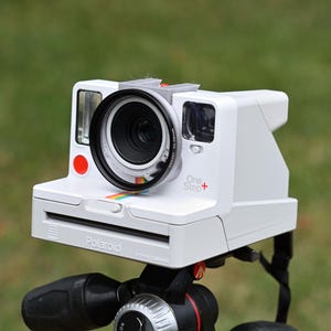 May include: A white Polaroid OneStep+ instant camera with a black lens and a red button. The camera is mounted on a black tripod with a red knob.