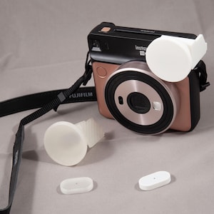 Peut inclure: Un appareil photo Instax Mini rose doré et noir avec un cache objectif blanc et une sangle. Deux petits accessoires blancs sont devant l'appareil photo. Le mot "FUJIFILM" est imprimé sur la sangle.