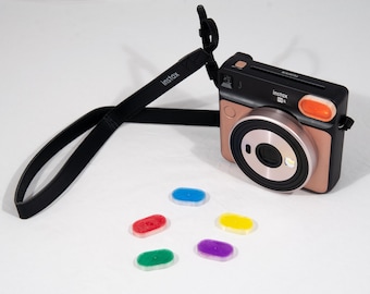 May include: A black and copper Instax Mini Evo instant camera with a black strap that says "Instax". The camera has an orange button and a silver lens. There are six colored lens filters: red, green, blue, yellow, and purple.