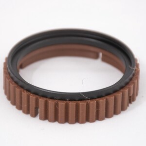 May include: A brown and black plastic ring with a gear-like design. The ring has a smooth black inner ring and a brown outer ring with a textured gear design.