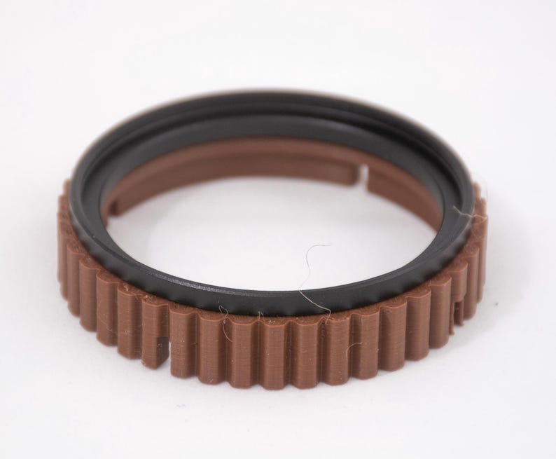 May include: A brown and black plastic ring with a gear-like design. The ring has a smooth black inner ring and a brown outer ring with a textured gear design.