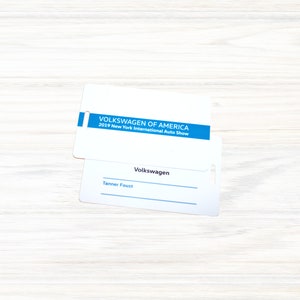 May include: White badge with blue text that reads "VOLKSWAGEN OF AMERICA 2019 New York International Auto Show". Below is a white badge with the text "Volkswagen" and a blank line for a name. The text "Tanner Foust" is written on the line.