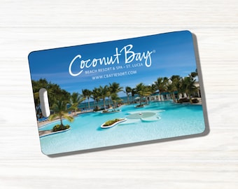 May include: A blue and white rectangular plastic sign with a handle. The sign has a photo of a swimming pool with palm trees and a blue sky. The text on the sign reads "Coconut Bay Beach Resort & Spa - St. Lucia www.cbayresort.com".