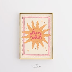 Op de afbeelding: Een ingelijste kunstafdruk met een zonne-illustratie in oranje en roze tinten. Het zonontwerp bevat de woorden "plein soleil" in cursief schrift. De print is ingelijst in een goudkleurige lijst.