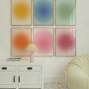 May include: Six framed abstract art prints with gradient colour backgrounds in yellow, blue, green, orange, pink, and purple. The prints are arranged in a grid on a white wall. A white cabinet with two drawers and two doors is below the prints. A lamp with a white shade and a pink base is on the cabinet. A light green chair is in the bottom right corner of the image.