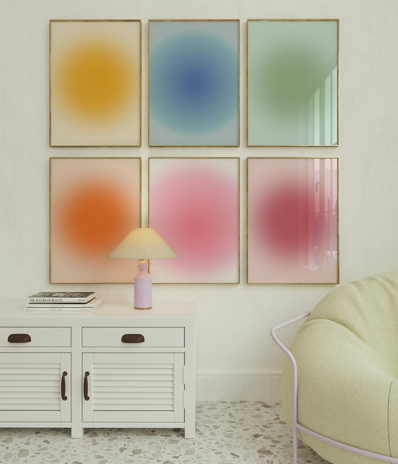 May include: Six framed abstract art prints with gradient colour backgrounds in yellow, blue, green, orange, pink, and purple. The prints are arranged in a grid on a white wall. A white cabinet with two drawers and two doors is below the prints. A lamp with a white shade and a pink base is on the cabinet. A light green chair is in the bottom right corner of the image.
