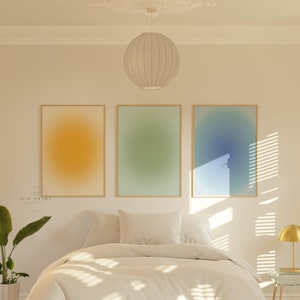 May include: Three framed abstract art prints with gradient colour backgrounds. The prints are in a bedroom setting above a white bed with white bedding and pillows. The prints are in a light yellow, light green, and light blue colour scheme.