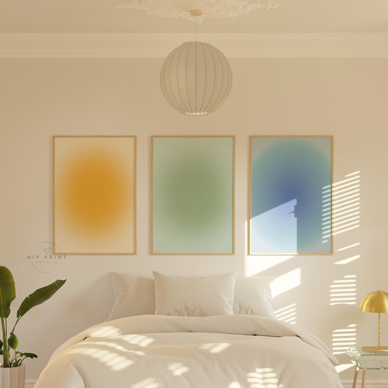 May include: Three framed abstract art prints with gradient colour backgrounds. The prints are in a bedroom setting above a white bed with white bedding and pillows. The prints are in a light yellow, light green, and light blue colour scheme.
