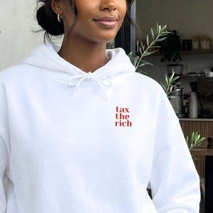 Puede incluir: Una sudadera con capucha blanca con el texto rojo "tax the rich". La sudadera tiene un bolsillo delantero y una capucha con cordón. La persona lleva vaqueros azul claro.
