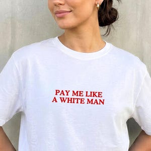 Könnte beinhalten: Weißes T-Shirt mit dem roten Text "PAY ME LIKE A WHITE MAN". Das Shirt hat kurze Ärmel und einen Rundhalsausschnitt. Das Model trägt hellblaue Jeans.