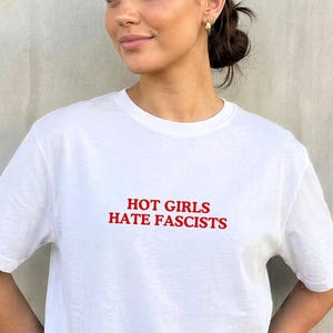 Puede incluir: Camiseta blanca con el texto rojo "HOT GIRLS HATE FASCISTS". La camiseta tiene mangas cortas y cuello redondo. La modelo lleva vaqueros azul claro.