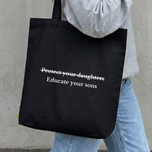 Könnte beinhalten: Eine schwarze Tragetasche mit weißem Text, der Protect your daughters Educate your sons lautet. Die Tasche hat zwei schwarze Schulterriemen und eine rechteckige Form, gefertigt aus einem einfarbigen Stoff mit einem schlichten Design.