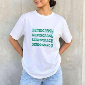 Op de afbeelding: Wit T-shirt met het woord "DEMOCRACY" vier keer groen gedrukt. Het shirt heeft korte mouwen en een ronde hals. Het model draagt een lichtblauwe jeans.