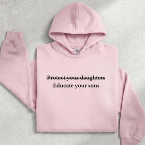 Op de afbeelding: Een lichtroze hoodie met lange mouwen en een voorzak. Op de borst staat de zwarte tekst "Protect your daughters" met een horizontale lijn door de woorden, gevolgd door de tekst "Educate your sons" die eronder is geplaatst.