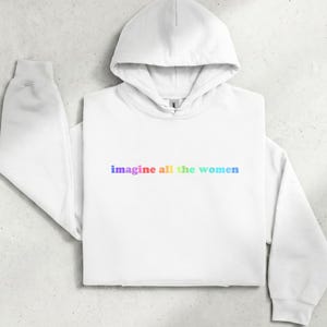 Könnte beinhalten: Ein weißer Kapuzenpullover mit langen Ärmeln und dem Schriftzug "imagine all the women", der in einem Regenbogenfarbverlauf auf die Brust gedruckt ist. Der Hoodie verfügt über eine Kapuze mit Kordelzug, lange Ärmel mit Bündchen und eine Kängurutasche.