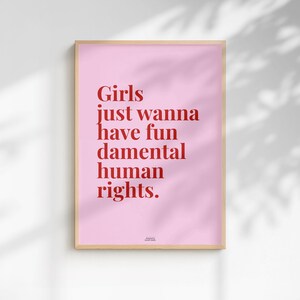 Könnte beinhalten: Ein rechteckiger Wandkunstdruck in einem hellen Holzrahmen mit einem hellrosa Hintergrund und fettgedrucktem rotem Text, der lautet: Girls just wanna have fundamental human rights. Das Design ist mittig platziert mit einer kleinen Quellenangabe unten.