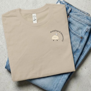 Op de afbeelding: Een beige t-shirt met ronde hals en een kleine afbeelding van een glimlachende dumpling met een koksmuts, met de tekst "dumplings & democracy" er in een boog boven. Het shirt is opgevouwen en ligt op een blauwe spijkerbroek.