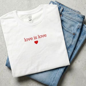 Könnte beinhalten: Ein weißes T-Shirt mit Rundhalsausschnitt, das den roten Schriftzug love is love und ein kleines rotes Herzsymbol unter dem Text zeigt, platziert neben einer blauen Jeans. Das T-Shirt hat ein kurzes Ärmeldesign und einen einfachen Halsausschnitt.