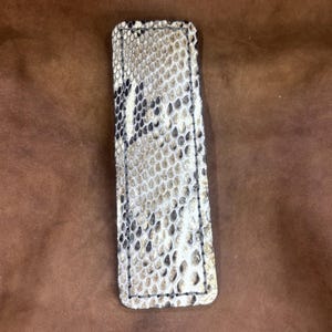 Op de afbeelding: Een rechthoekig leren item met een slangenpatroon in tinten wit, beige en zwart. De randen zijn gestikt met zwarte draad. Het item staat tegen een bruine achtergrond.