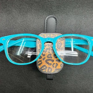 May include: Bright turquoise plastic eyeglasses resting on a vertical magnetic holder featuring a brown and black leopard print pattern. The holder has a dark gray leather-like tab at the top and a small black fabric loop at the bottom edge.
