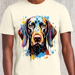 以下が含まれることがあります： クリーム色のTシャツに、犬の顔のカラフルな抽象画が描かれています。犬は茶色の毛に黒い斑点があります。絵には、青、黄、オレンジ、赤の塗料の飛沫が描かれています。