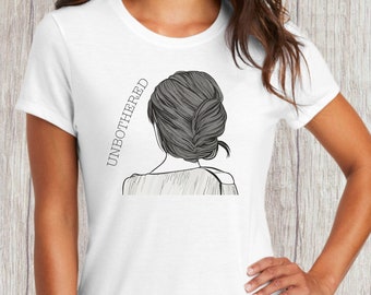 Peut inclure: T-shirt blanc avec un dessin au trait noir et blanc d'une femme avec les cheveux tirés en arrière en chignon. Le texte "UNBOTHERED" est écrit verticalement sur le côté gauche de l'image.