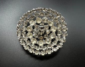 May include: A silver-toned, circular brooch or button, densely covered with clear, sparkling rhinestones. The design features concentric circles of stones, with a central, raised element. The piece is set against a dark background.