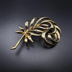 May include: A gold-toned brooch in a floral design. The brooch features a stylized leaf and swirl design, with a textured surface. The brooch is set against a dark background.